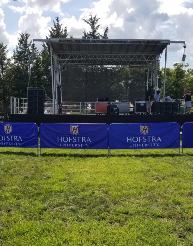 NYC portable event stage and bandshell rentals. portable, rentable event stage with blue banners on it, An event stage customizable and portable, with an open top and a person walking on the grass to rent in Long Island and Tri state area from Allseasonsstagerentals.com